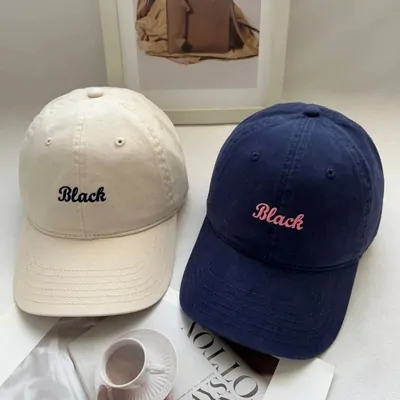 Cream and navy baseball caps with script embroidery, on a desk beside a magazine